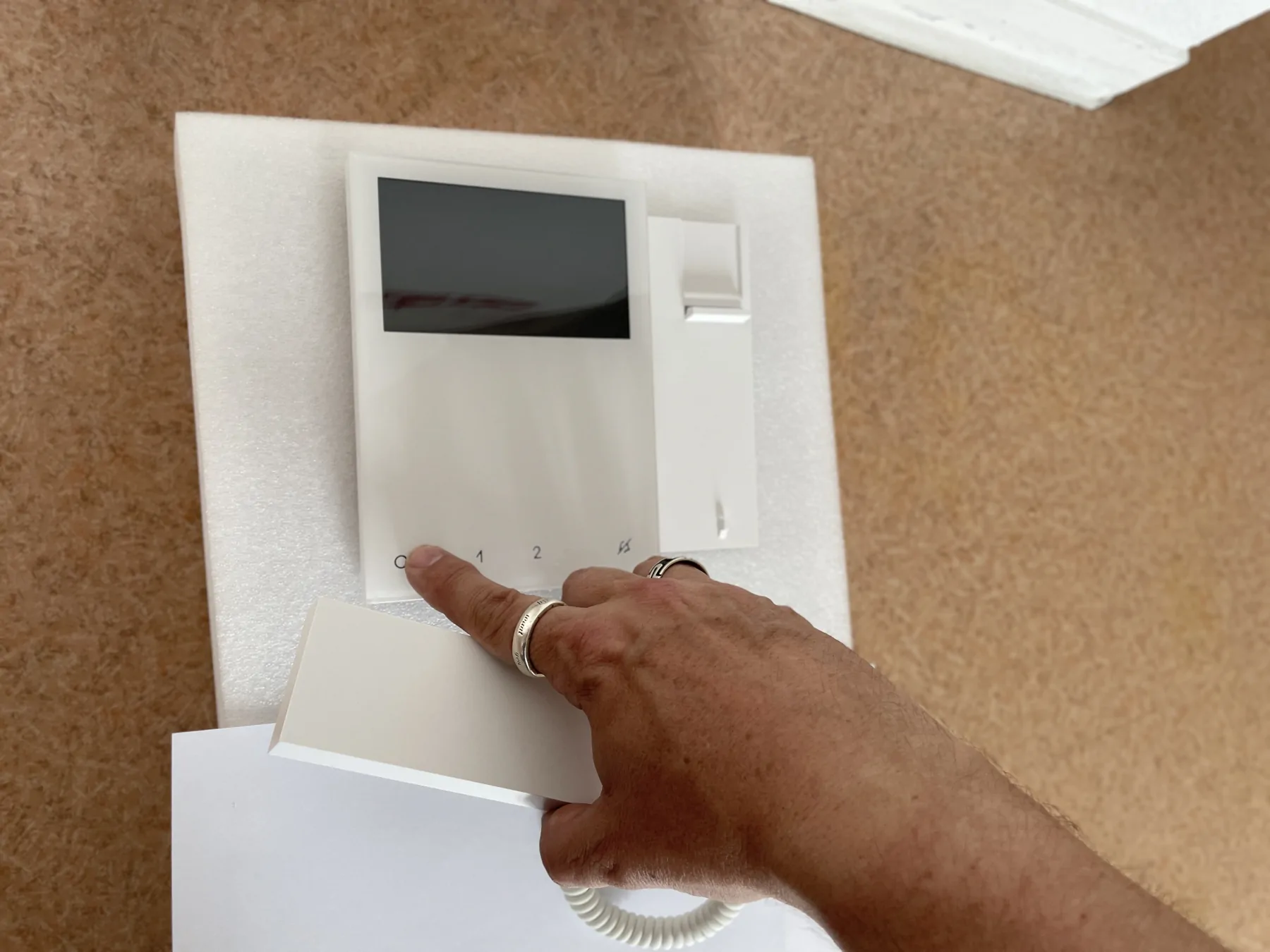 Une main appuie sur un bouton d’un visiophone mural blanc.